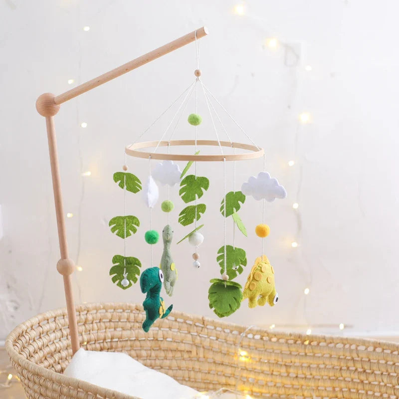 Wooden dinosaur crib mobile with soft felt dinosaur toys and green leaf decorations hanging above a wicker baby bassinet.