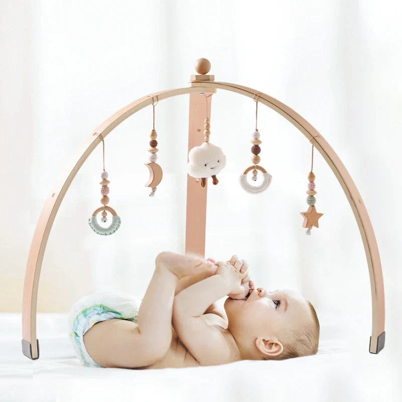 Wooden baby play gym with hanging sensory toys including moon, cloud, star, and rings over a baby lying on a white surface.