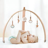 Wooden baby play gym with hanging sensory toys including moon, cloud, star, and rings over a baby lying on a white surface.